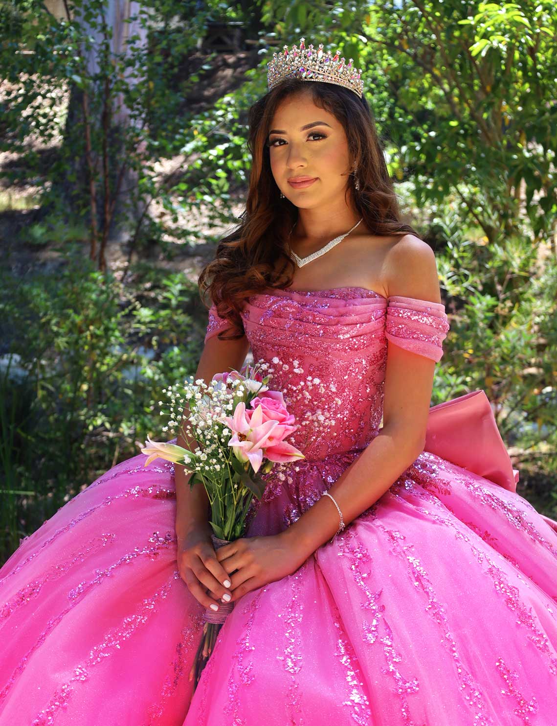 Young woman in a pink off-the-shoulder ball gown with a jeweled crown, holding a pink bouquet outdoors.